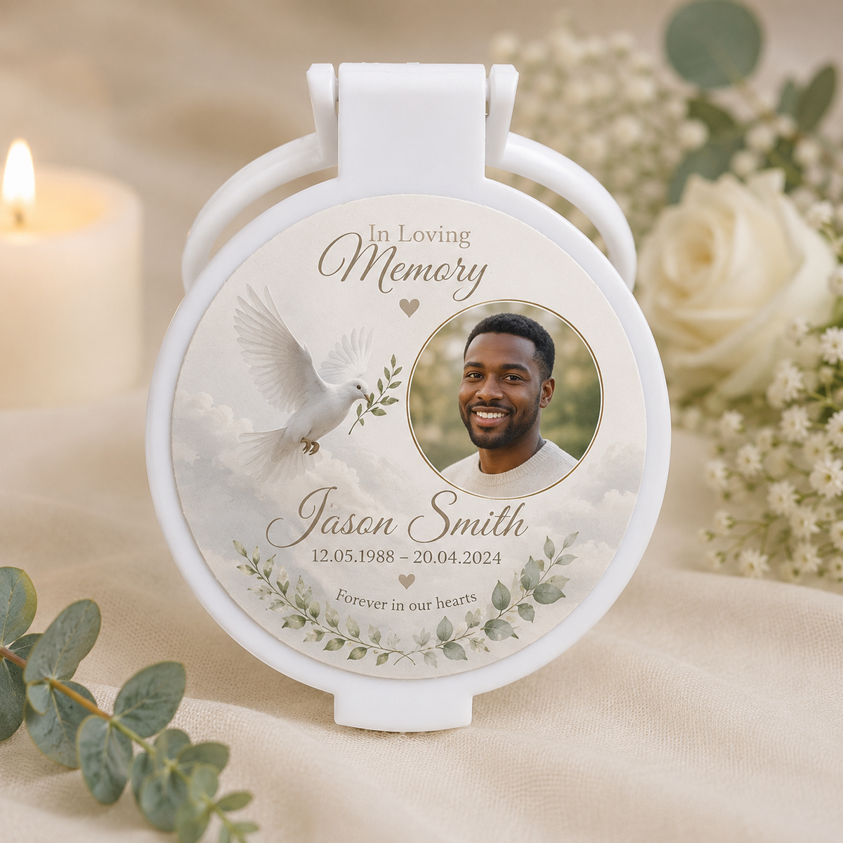 A white circular folding keepsake mirror with a printed memorial design, shown on a soft beige fabric with a lit candle, white roses, and greenery in the background. The printed design includes the text 'In Loving Memory', a photo of a smiling man, the name 'Jason Smith', dates, and floral details.