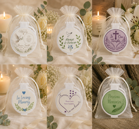 Six round memorial keepsake boxes in clear tulle drawstring bags, each with a different printed memorial label in white, purple, blue, green, and gray. The boxes are arranged in a 2x3 grid with soft-focus candles, flowers, and greenery in the background.