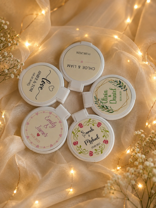 Five personalised wedding compact mirror favours in different designs displayed on elegant wedding fabric with warm fairy lights